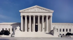 The image shows the U.S. Supreme Court building, featuring a symmetrical facade with tall columns and intricate sculptures, under a clear blue sky.