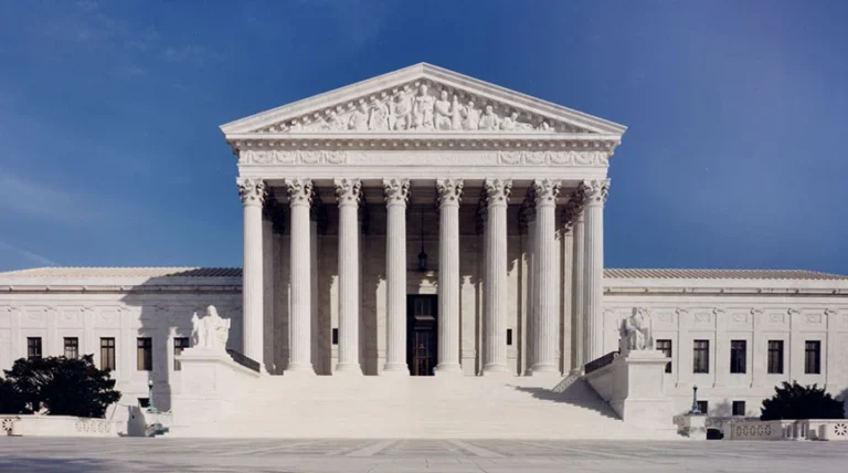 The image shows the U.S. Supreme Court building, featuring a symmetrical facade with tall columns and intricate sculptures, under a clear blue sky.