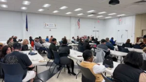 A diverse group of people sits at round tables in a large, bright meeting room with white walls. The atmosphere appears attentive and engaged.