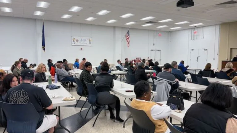 A diverse group of people sits at round tables in a large, bright meeting room with white walls. The atmosphere appears attentive and engaged.