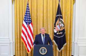 A man in a suit speaks at a podium with the U.S. presidential seal, flanked by the American flag and the presidential flag in a gold-curtained room.