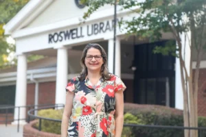A woman wearing glasses and a floral dress stands smiling in front of the Roswell Library entrance surrounded by trees.