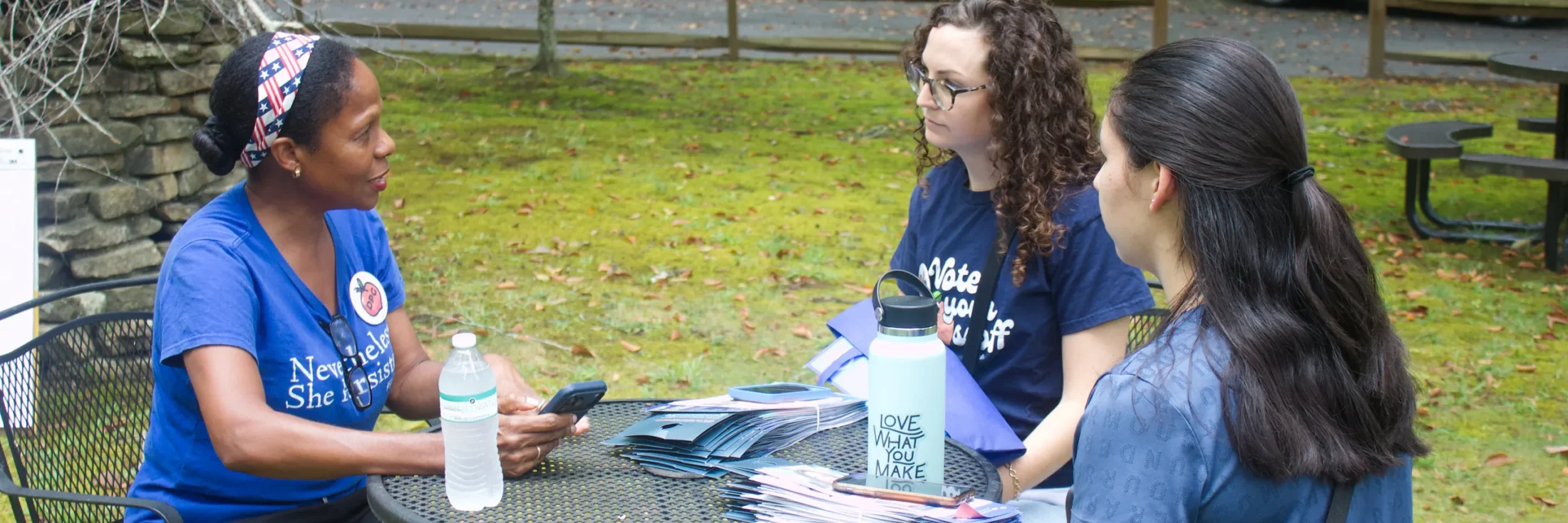 Three Fulton Democrats volunteers sitting around an outdoor table in conversation, with stacks of addressed voter mail between them.