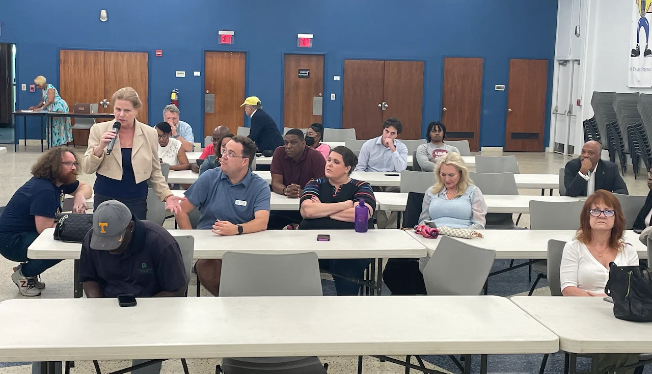 A committee member standing with a microphone addressing people seated at long tables during a Fulton County Democrats meeting.