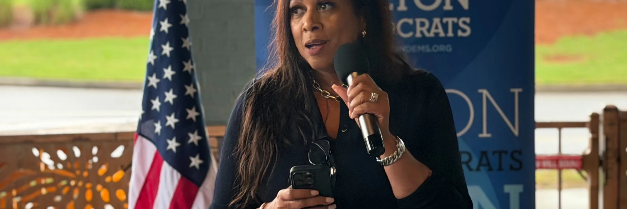 A speaker holding a microphone in front of a Fulton Democrats banner showing the fultondems.org address.