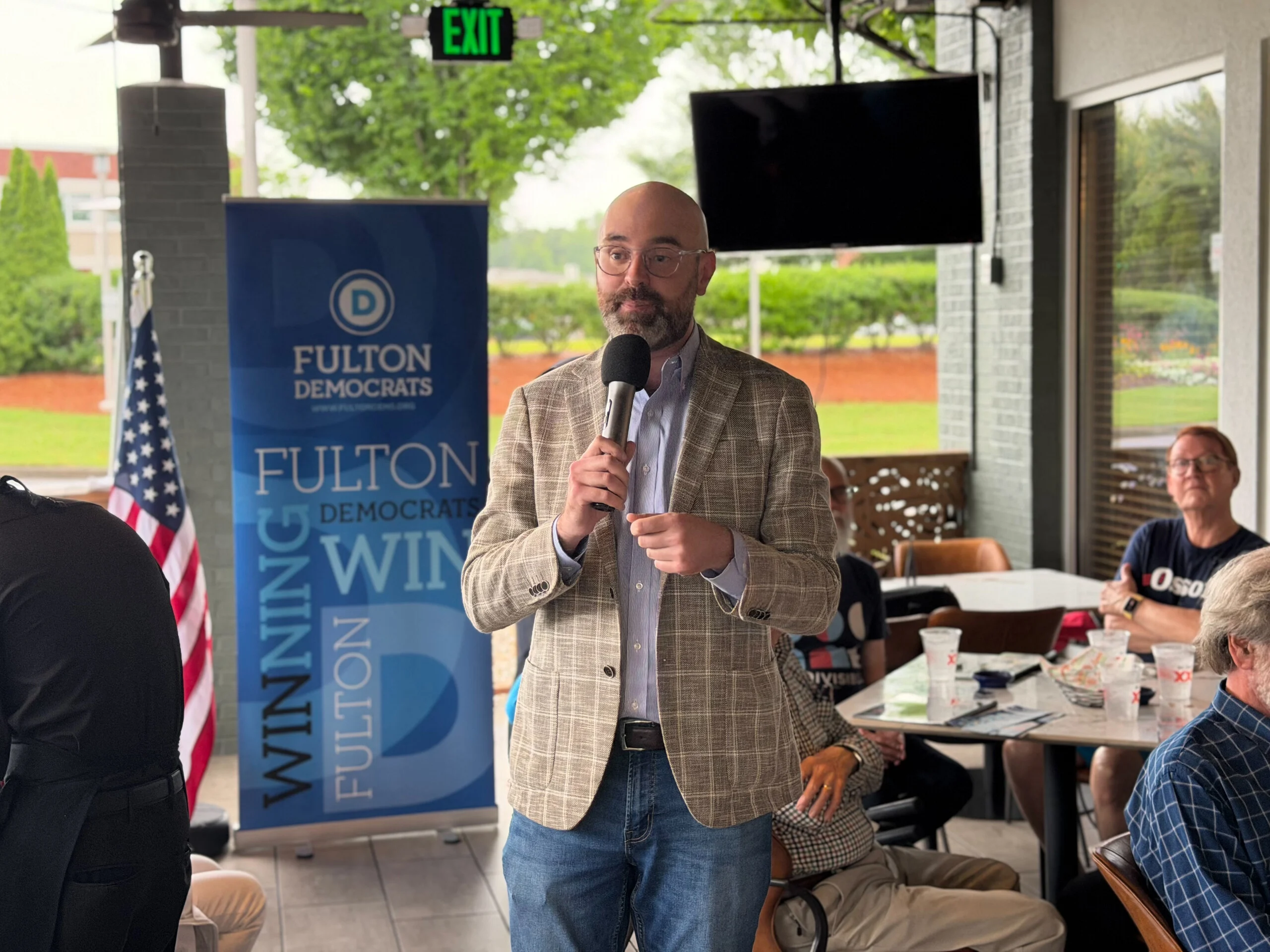 A speaker addresses attendees at a Fulton Democrats candidate forum, standing beside a "Fulton Democrats Winning Fulton" banner.